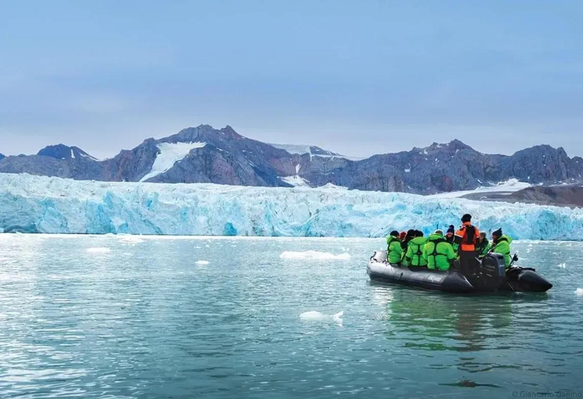 A boat with people in green jackets floats near a glacier and mountains.