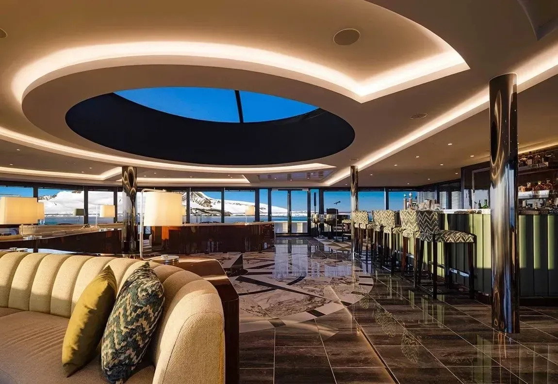 Lounge on a cruise ship with panoramic views, bar, skylight, plush seating, and a tiled floor.