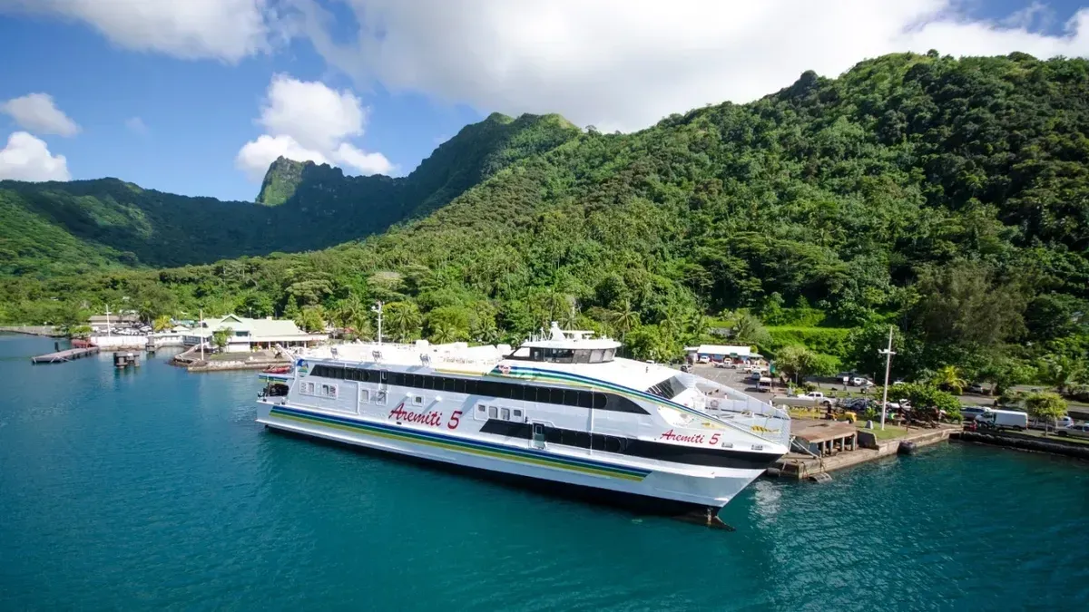 Ferry docked at pier with lush green mountains in the background and a blue sky.