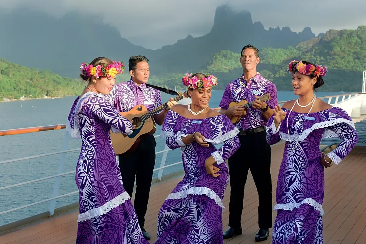 Five people in purple Polynesian attire playing music and dancing on a cruise ship deck, mountains in the background.