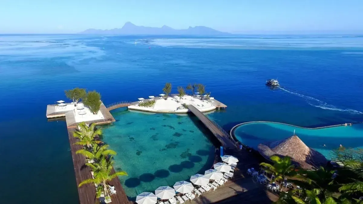 Aerial view of a tropical resort with turquoise water, a small island, and a pier, lush greenery, and blue sky.