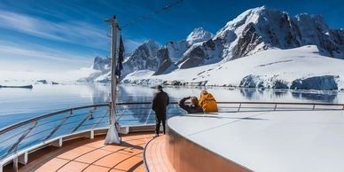 A person stands on a yacht deck, looking at snow-covered mountains and icy waters.