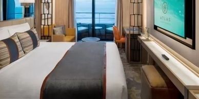 A modern cruise ship cabin with a bed, TV, balcony, and seating area.