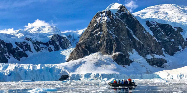 A boat with people navigates icy water in front of a snow-covered mountain range under a blue sky.