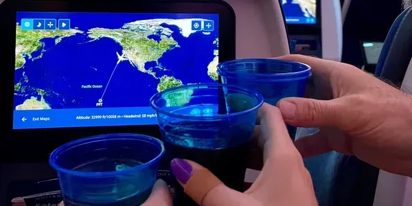 Hands holding blue cups on an airplane, flight map on the screen in the background.
