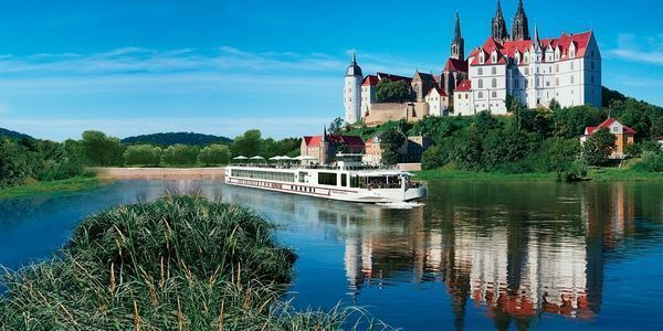 A white cruise ship on a blue river reflecting a castle on a hill under a blue sky.
