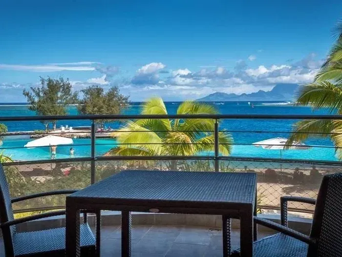 Balcony view of turquoise water, beach, distant mountains, and a table and chairs.