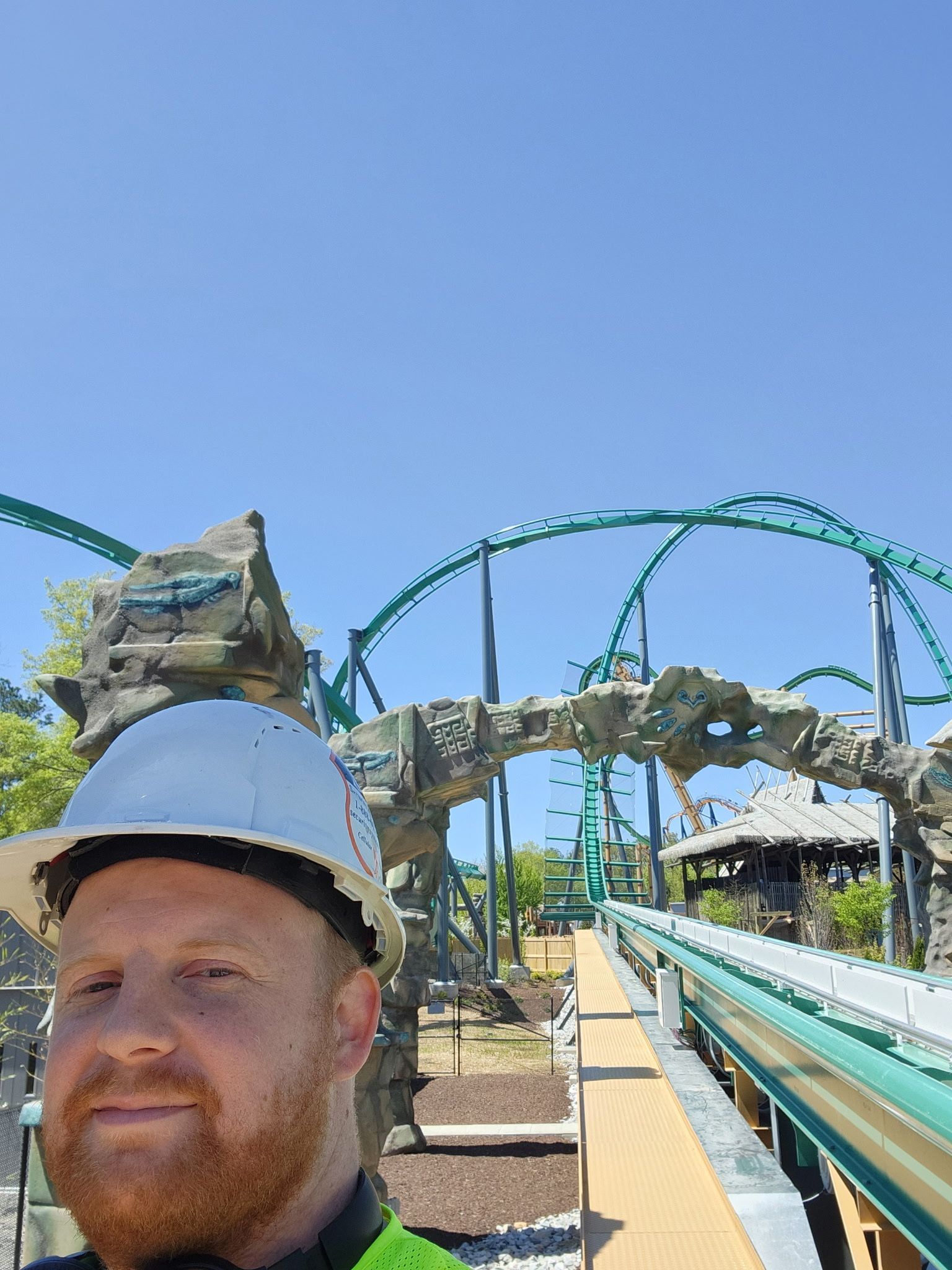 Man in hard hat smiles, roller coaster track and structure in background, blue sky.