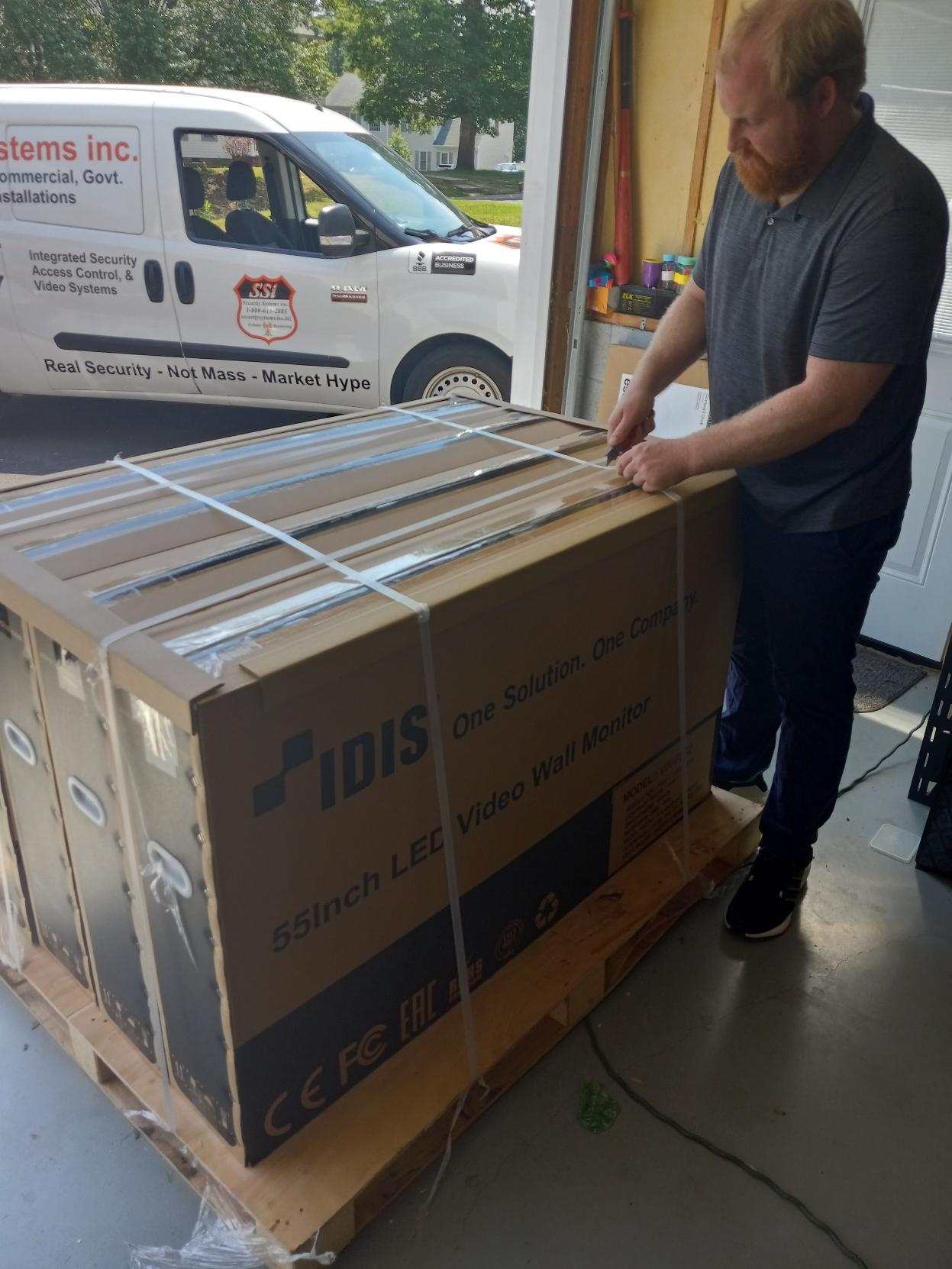Man opening large cardboard box on a pallet outside a garage. A white van is parked nearby.