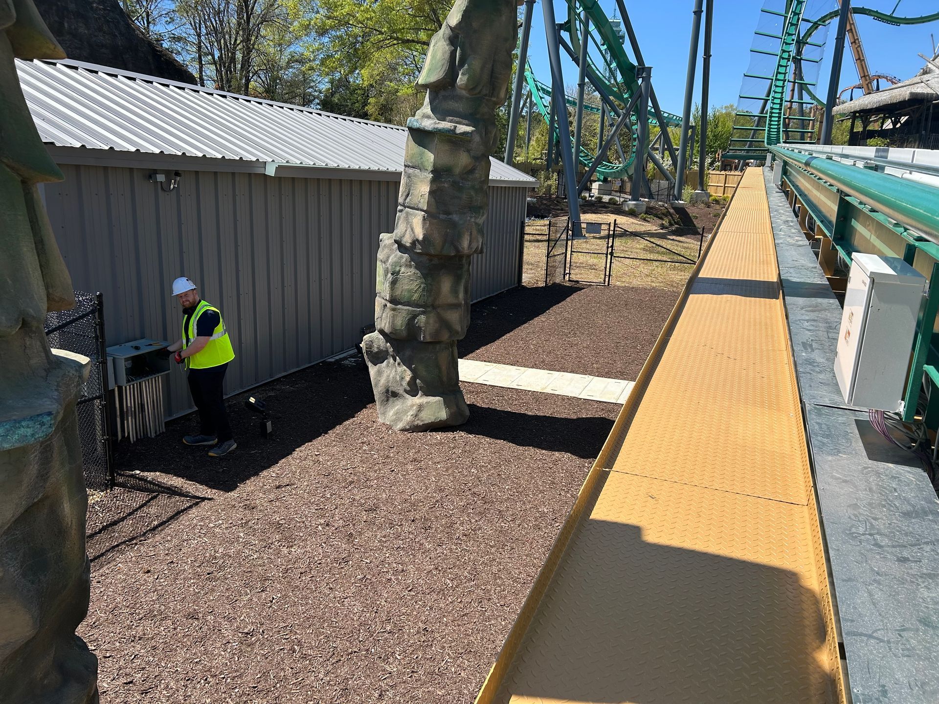 A person in a yellow vest near a rollercoaster track.  Gray shed and rocky ground. Bright sky.