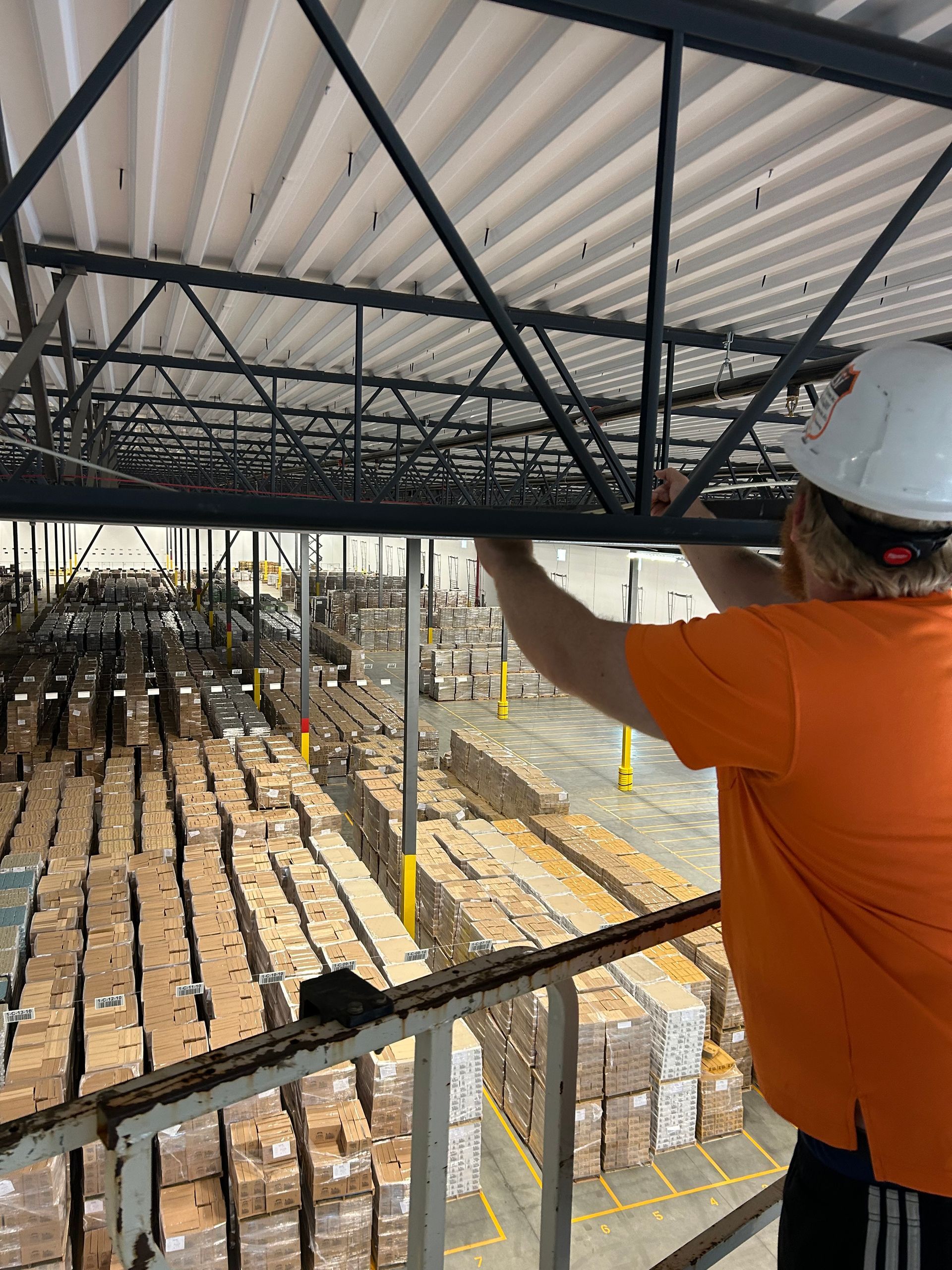 Person in orange shirt and hard hat working on metal beams above a warehouse filled with boxes.