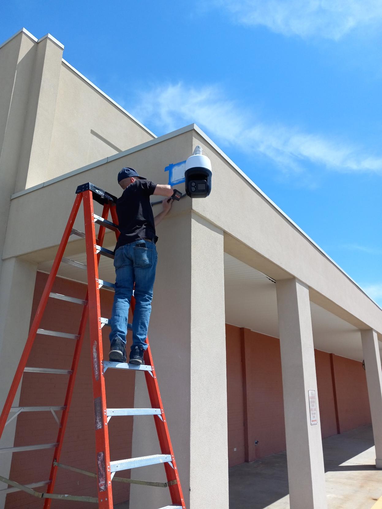 Person on a ladder installing a security camera on a building's corner.