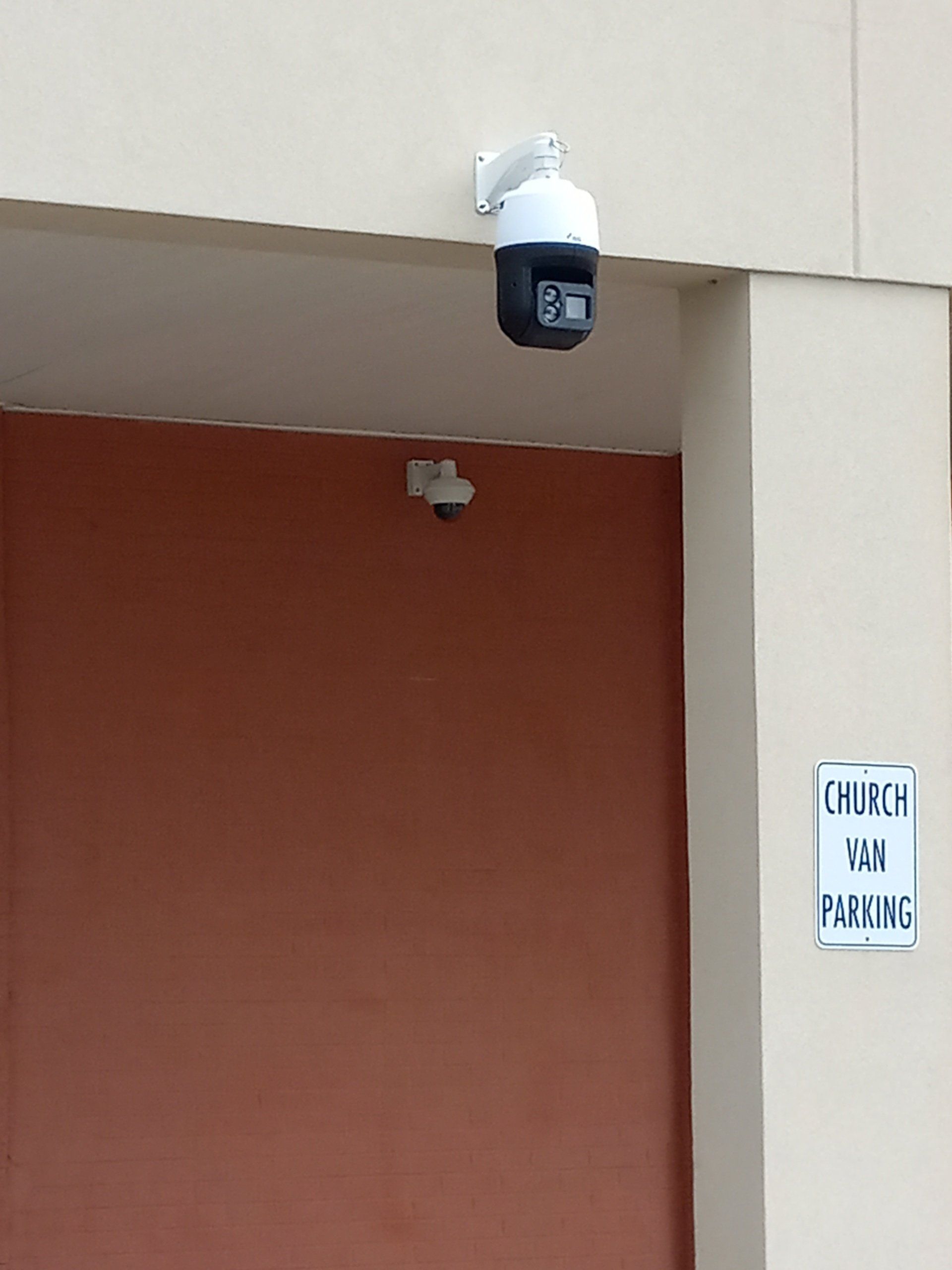Security cameras mounted on a building; one large, swiveling, and another smaller. Sign says 