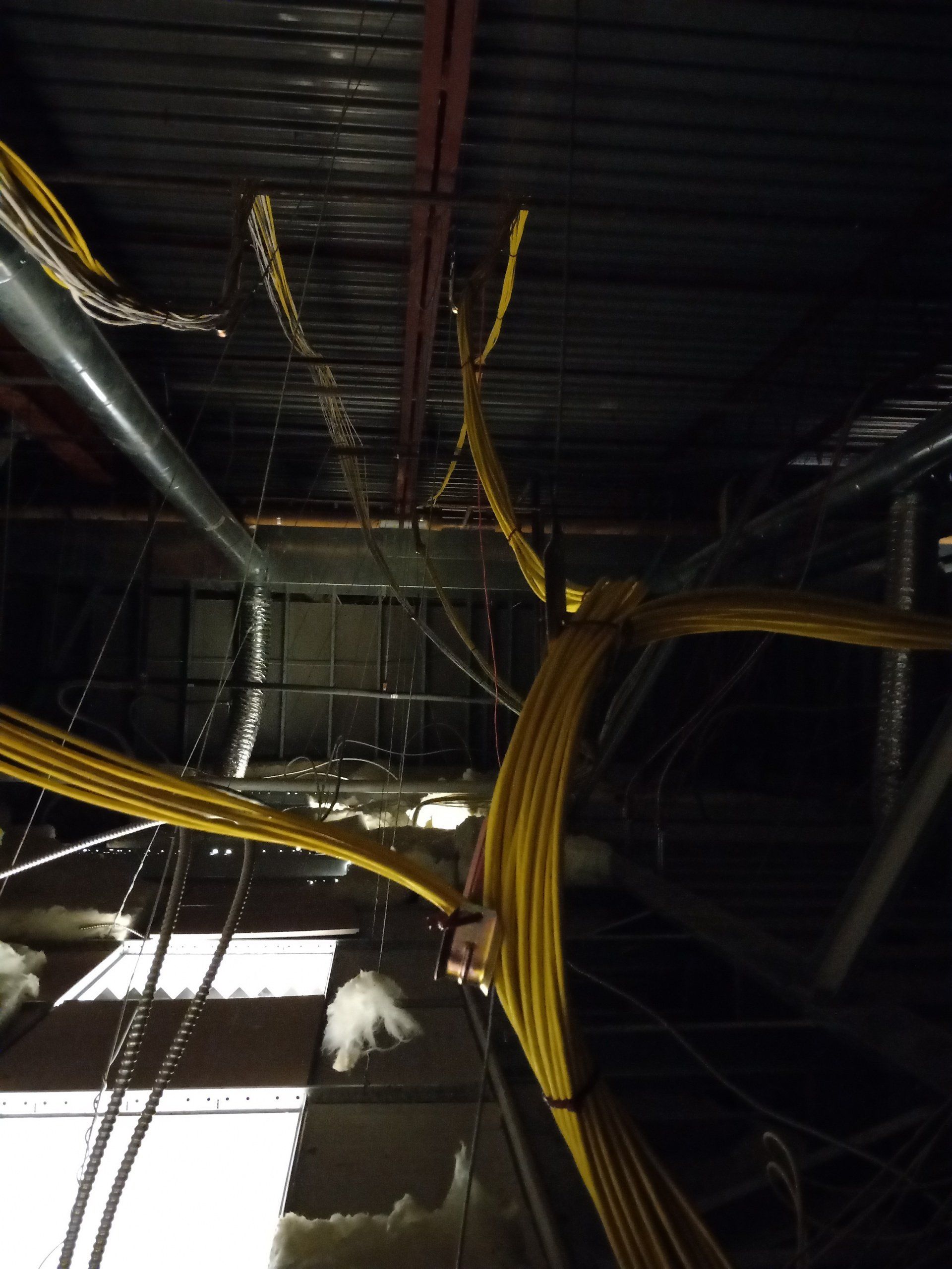 Overhead view of a construction site ceiling with numerous yellow electrical cables, ductwork, and exposed metal beams.