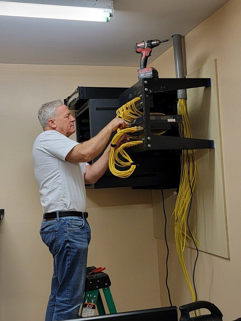 Man installing network cables on a wall-mounted rack, using a drill, inside a room. Yellow cables.