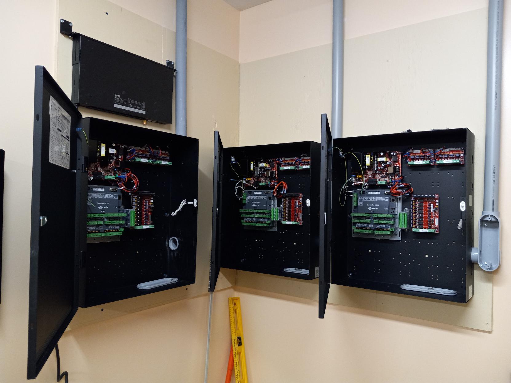 Three open black electrical control panels on a wall with wiring and circuit boards; gray conduit pipes above.