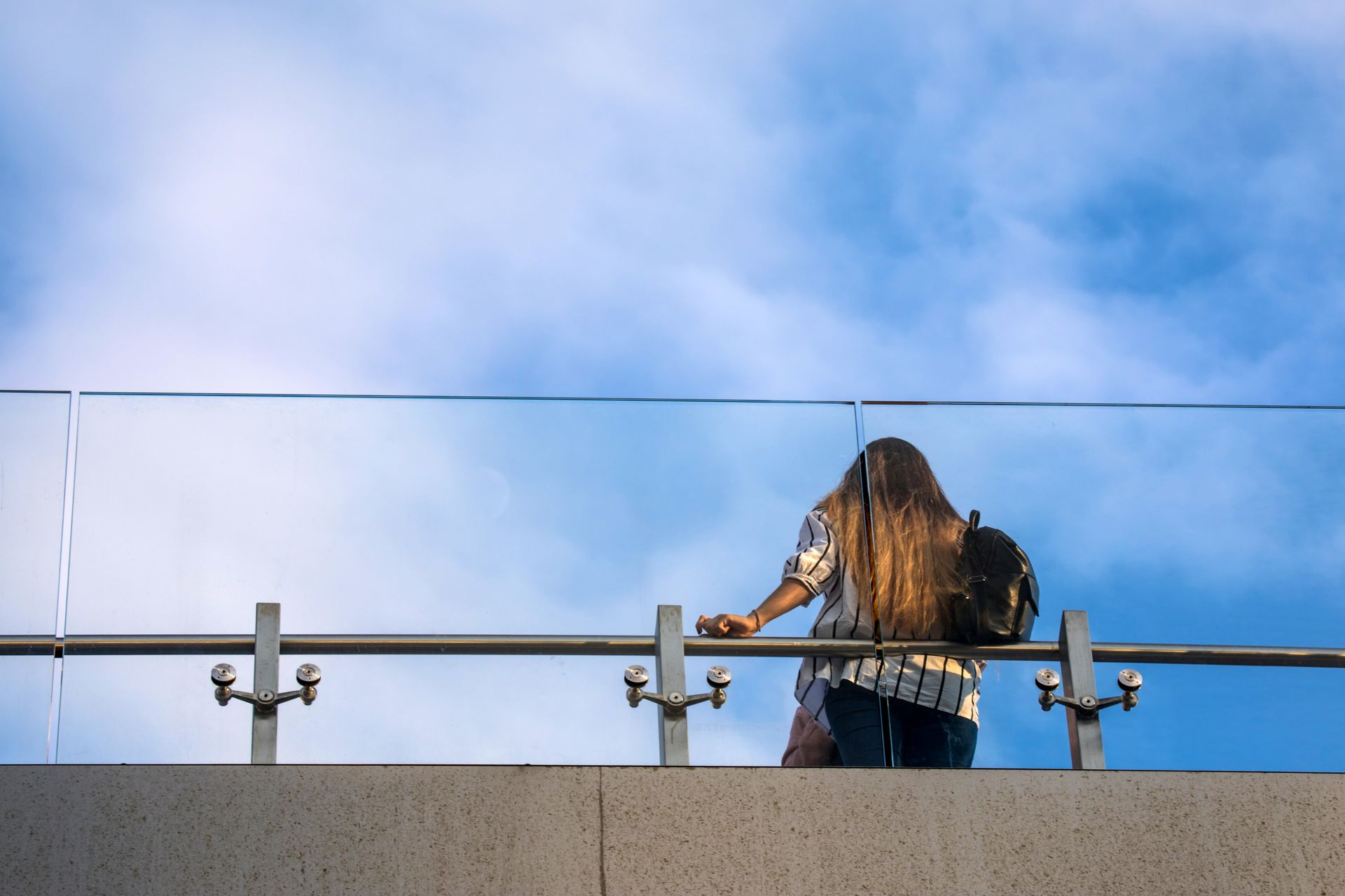 The rear side of a woman carrying a backpack standing on a glass deck balustrade.