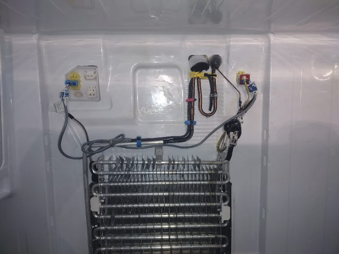 Inside a refrigerator, exposed cooling coils and components with wiring and tubing.