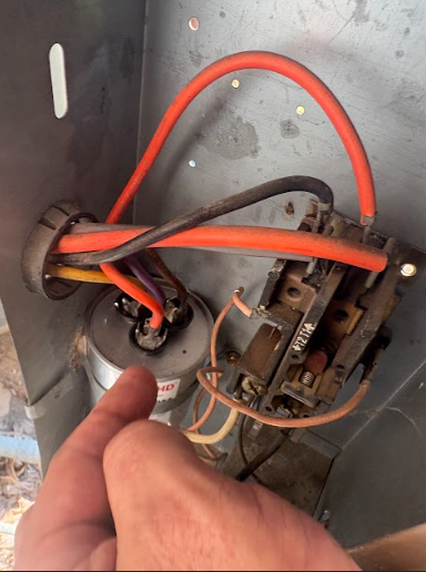 Hand pointing at air conditioning unit components. Orange, black, and yellow wires connect to a capacitor and a relay switch.