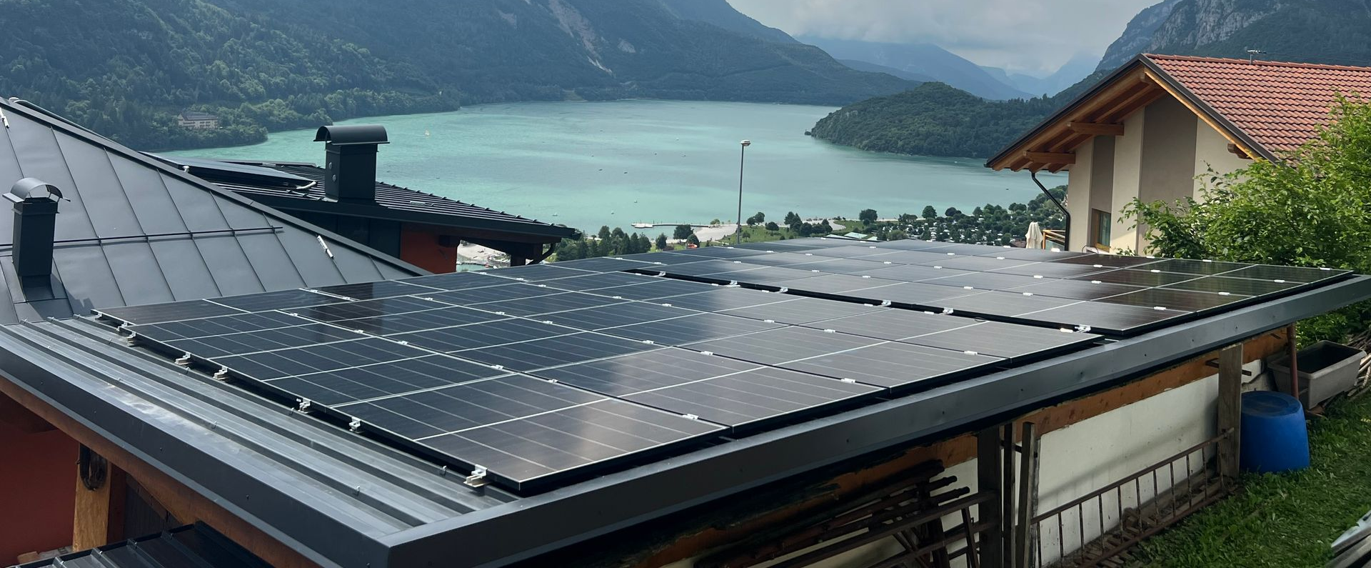 Pannelli solari su un tetto metallico con vista su un lago e sulle montagne, sotto un cielo nuvoloso.