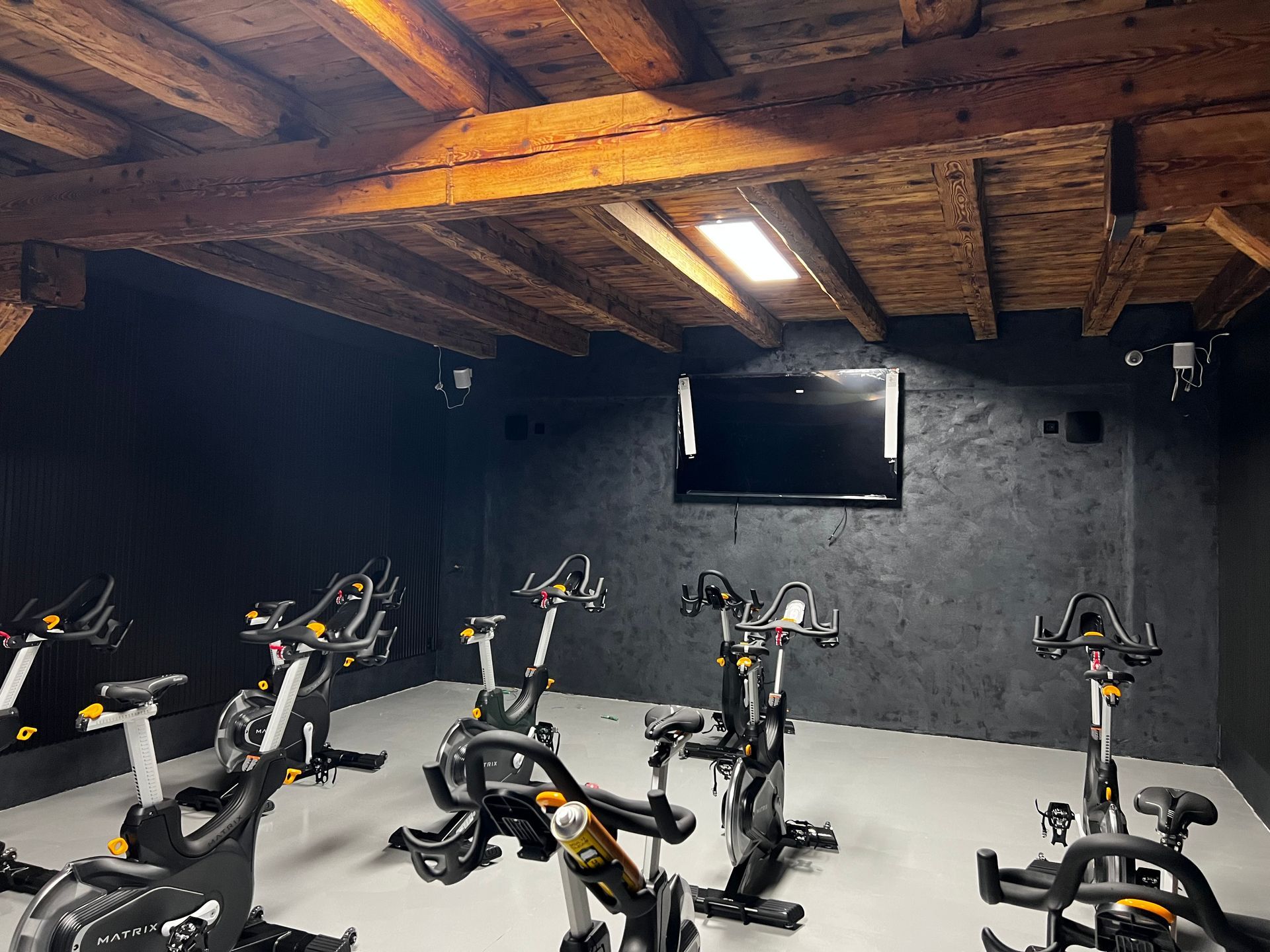 Sala per ciclismo indoor con diverse cyclette stazionarie su un pavimento grigio, pareti nere e soffitto con travi in ​​legno.