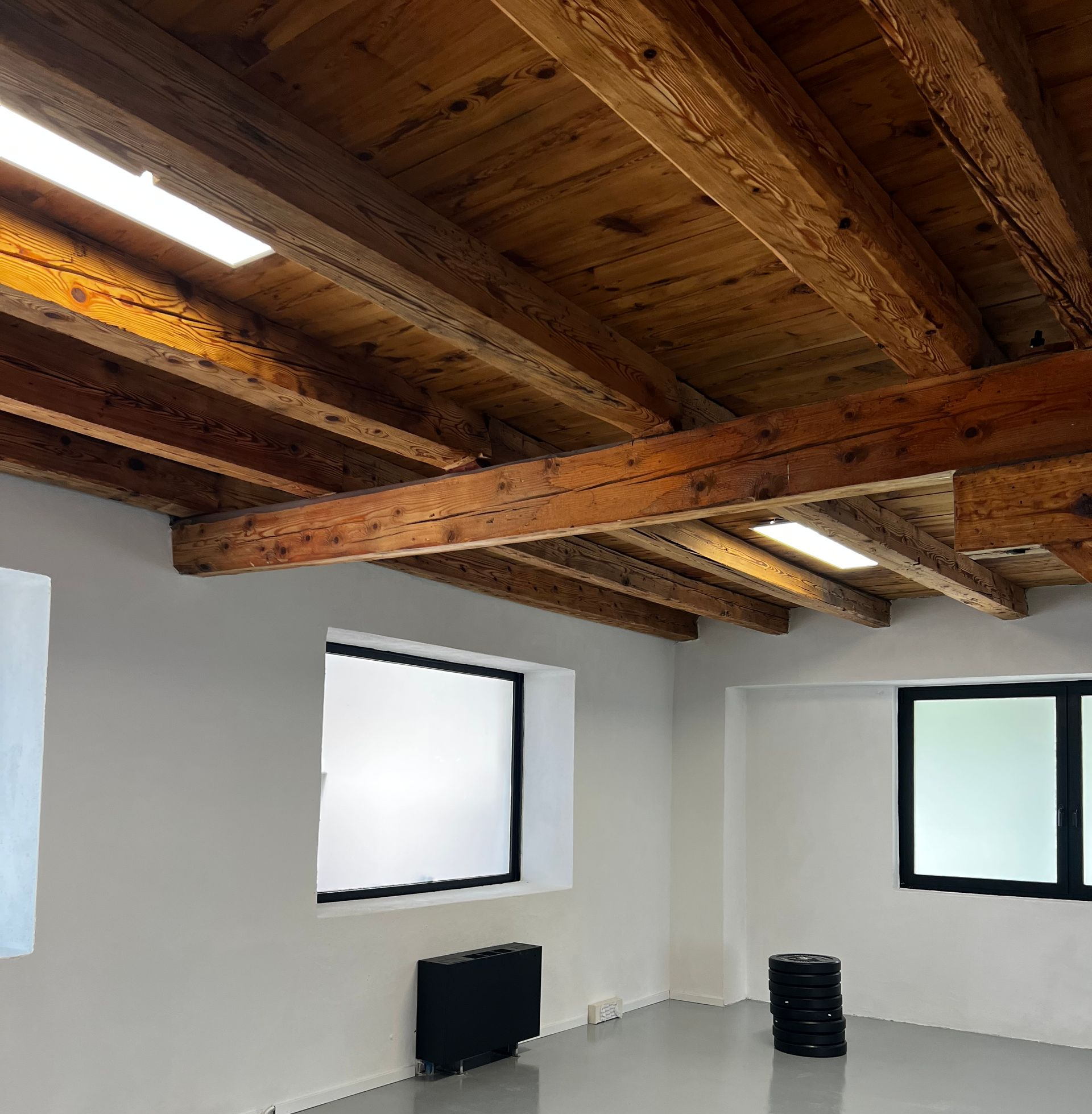 Una sala fitness con travi in ​​legno a vista, pareti bianche e pile di dischi per bilanciere neri su un pavimento grigio.