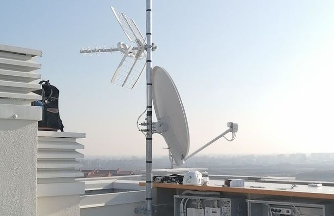 Una parabola satellitare bianca e un'antenna televisiva montate su un palo in cima a un tetto, stagliano un cielo limpido e pallido contro lo sfondo.