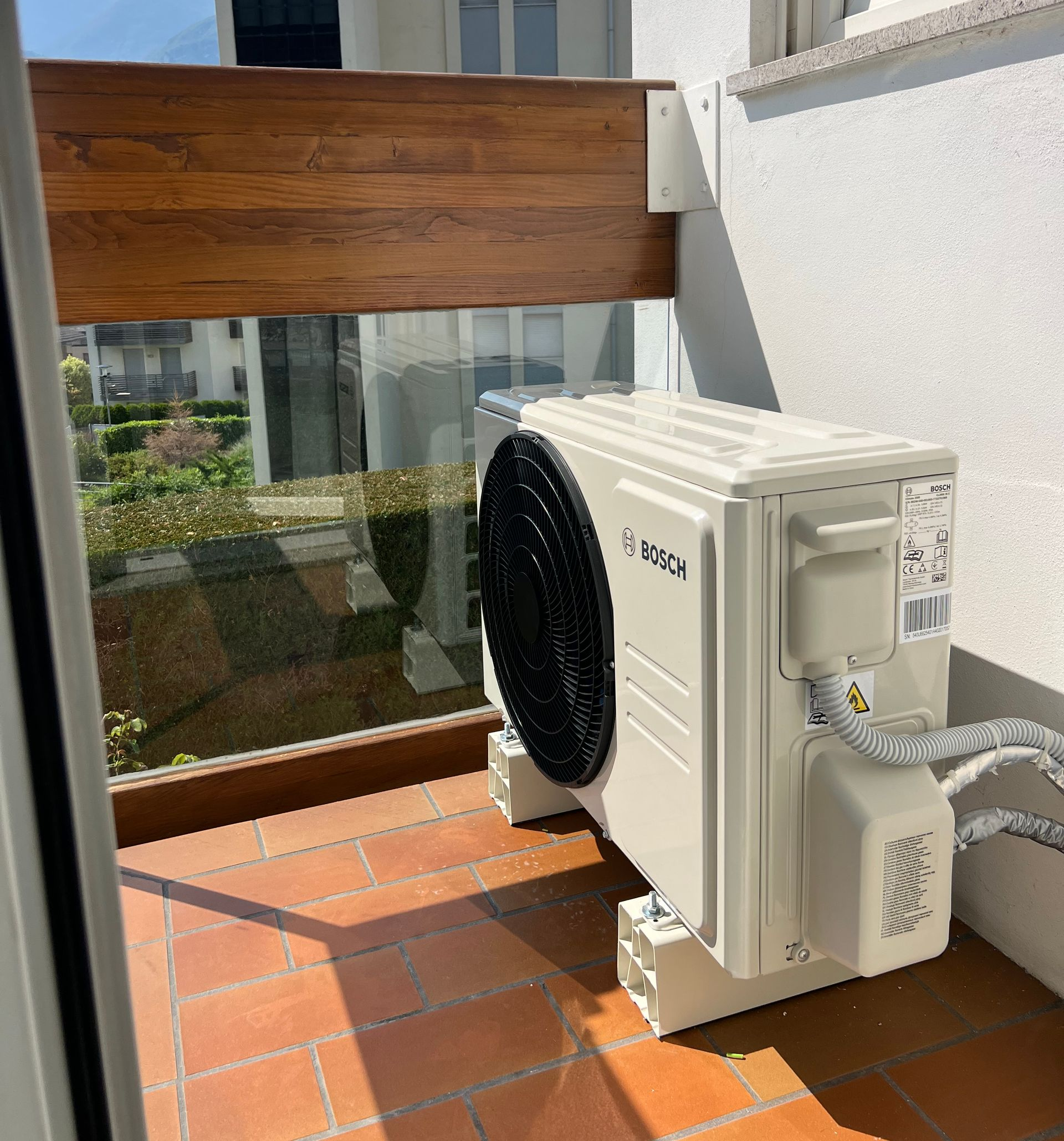 Un'unità esterna di un condizionatore d'aria Bosch di colore beige è installata su un balcone con piastrelle in terracotta e ringhiera in legno e vetro.