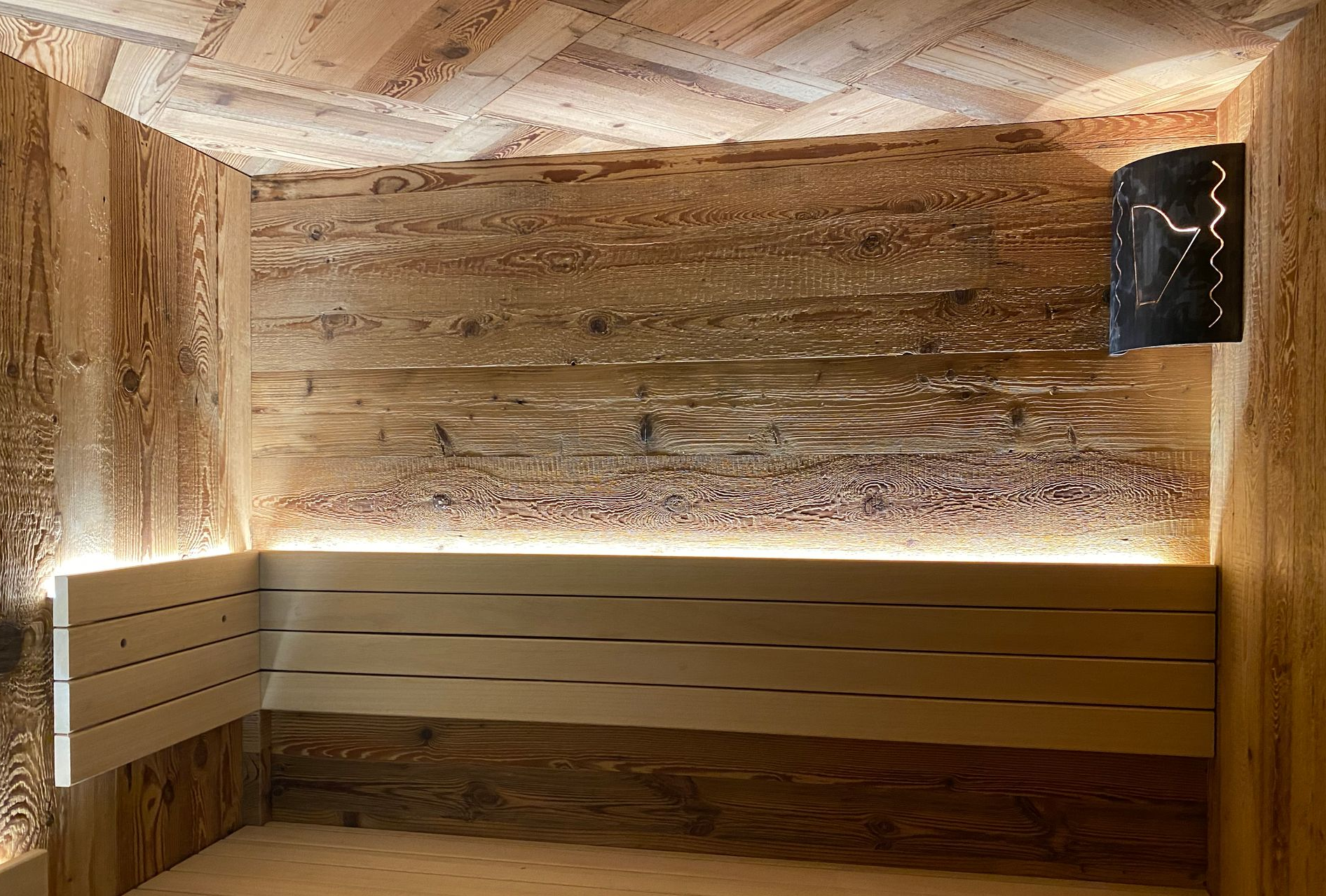 In una cantina, tra scaffali pieni di bottiglie di vino e un soffitto a doghe di legno, si trova un lungo tavolo di legno con sedie verdi.