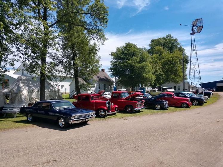 A row of classic cars parked on a gravel lot near trees, a white farmhouse, and a tall metal windmill.