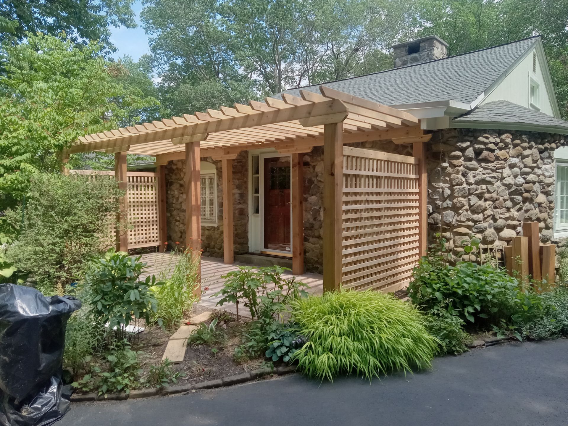 Wooden pergola over stone house entrance with lattice and surrounding greenery.