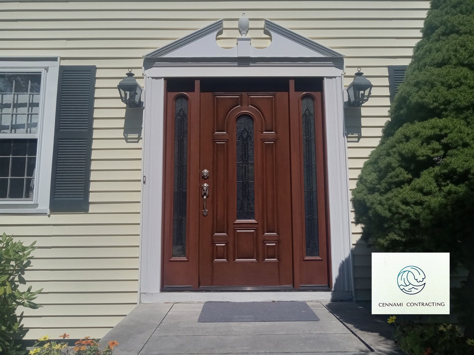 Brown front door with sidelights, under a decorative pediment. Beige siding and bushes.