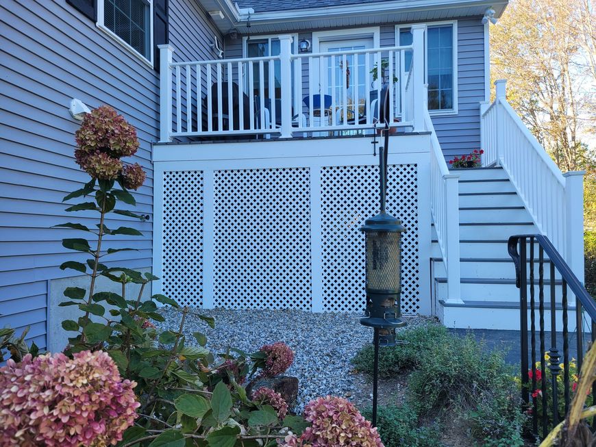 White deck with lattice skirting, stairs, and railing, next to a blue house with flowers and a bird feeder.