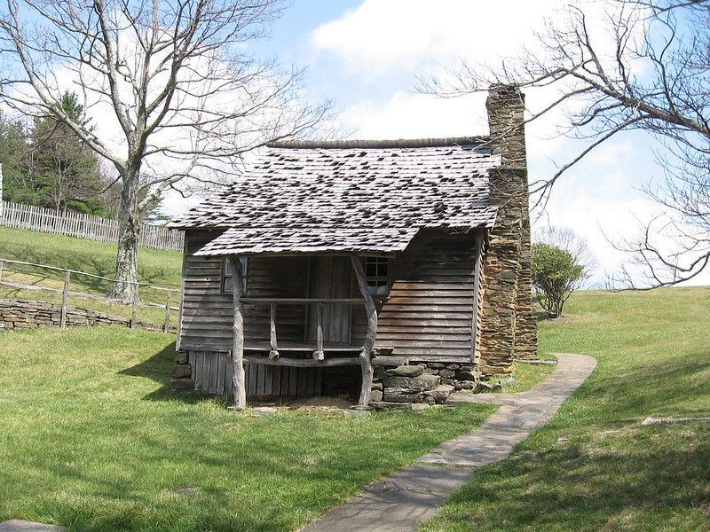 An old wooden cabin with a porch and chimney is sitting in the middle of a grassy field in the smoky mountains