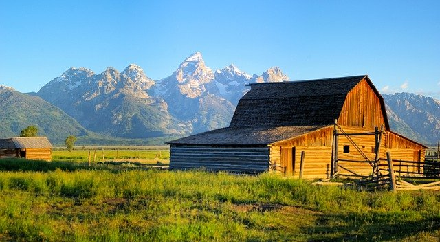 A historic barn is sitting in the middle of a grassy field with mountains in the background in Grand Teton NP, Wyoming