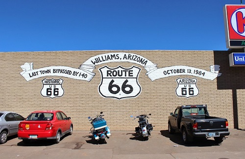 Cars and motorcycles are parked in front of a route 66 sign in Williams