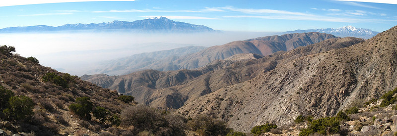 A panoramic view of a mountain range i Joshua Tree NP with trees and clouds.