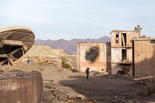 A man is walking in front of an abandoned building in Joshua Tree NP with mountains in the background.