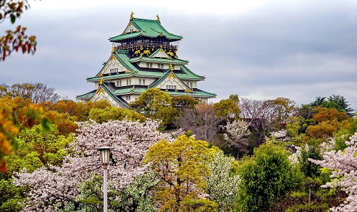 A large castle is surrounded by trees and flowers in a park in Osaka