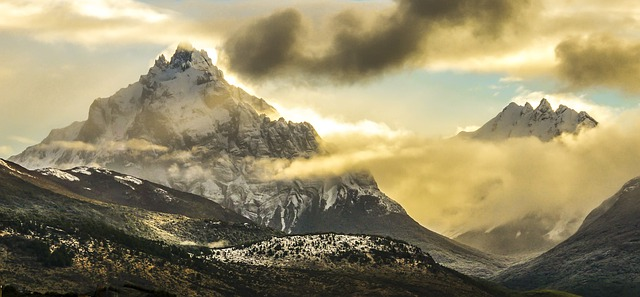 A mountain covered in snow and clouds with the sun shining through the clouds.