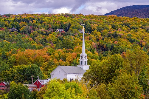 A white church is surrounded by trees in the middle of a forest in Stowe