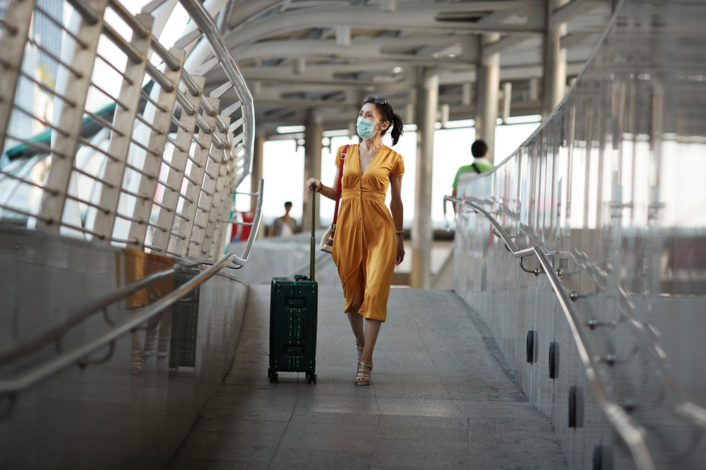 A woman wearing a mask is walking down a bridge with a suitcase.