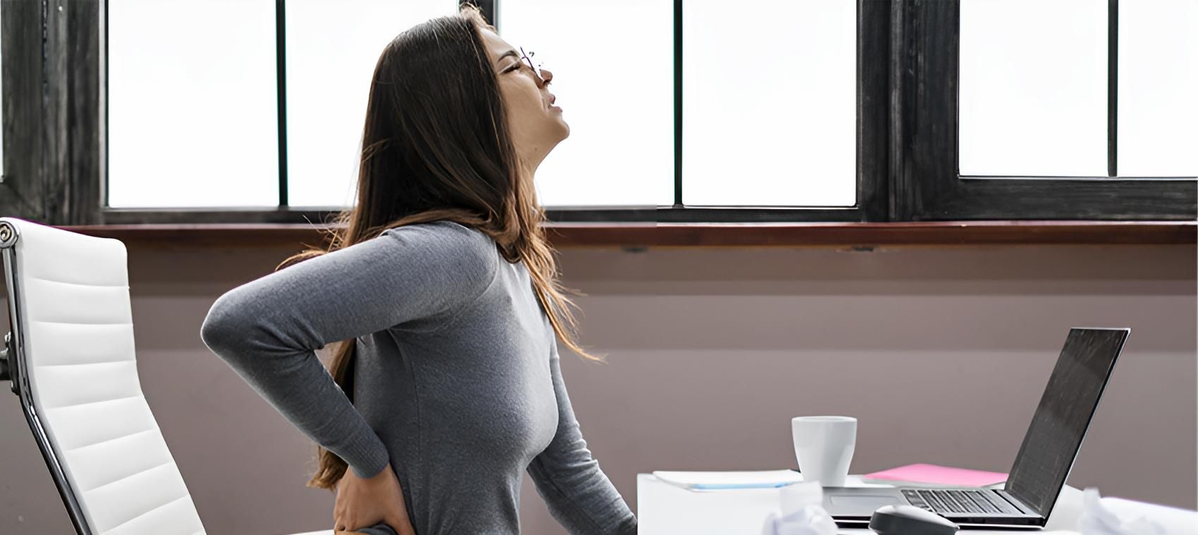 A Woman is Sitting at a Desk With a Laptop and Holding Her Back — Back Care Gold Coast in Nerang, QL