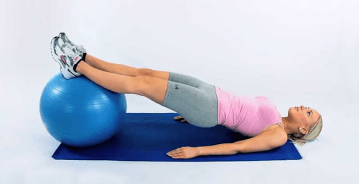 Uma mulher está deitada em uma esteira com as pernas apoiadas em uma bola de exercícios azul.