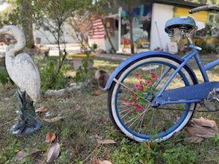 Blue bicycle with white tires, flowers in rear wheel, next to a white bird statue in a yard.
