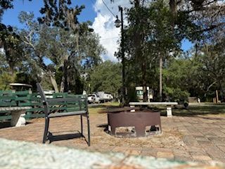 Park setting with a fire pit, benches, and trees under a blue sky.