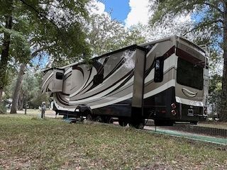 Large RV with extended slide-outs parked on a grassy lot, surrounded by trees.