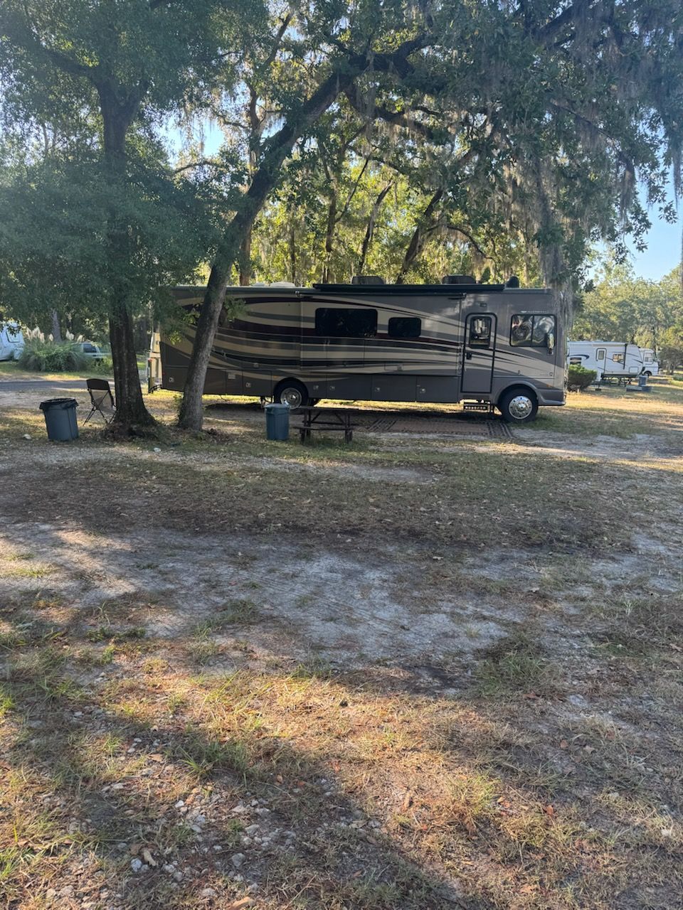 RV parked on a gravel lot under trees with Spanish moss.