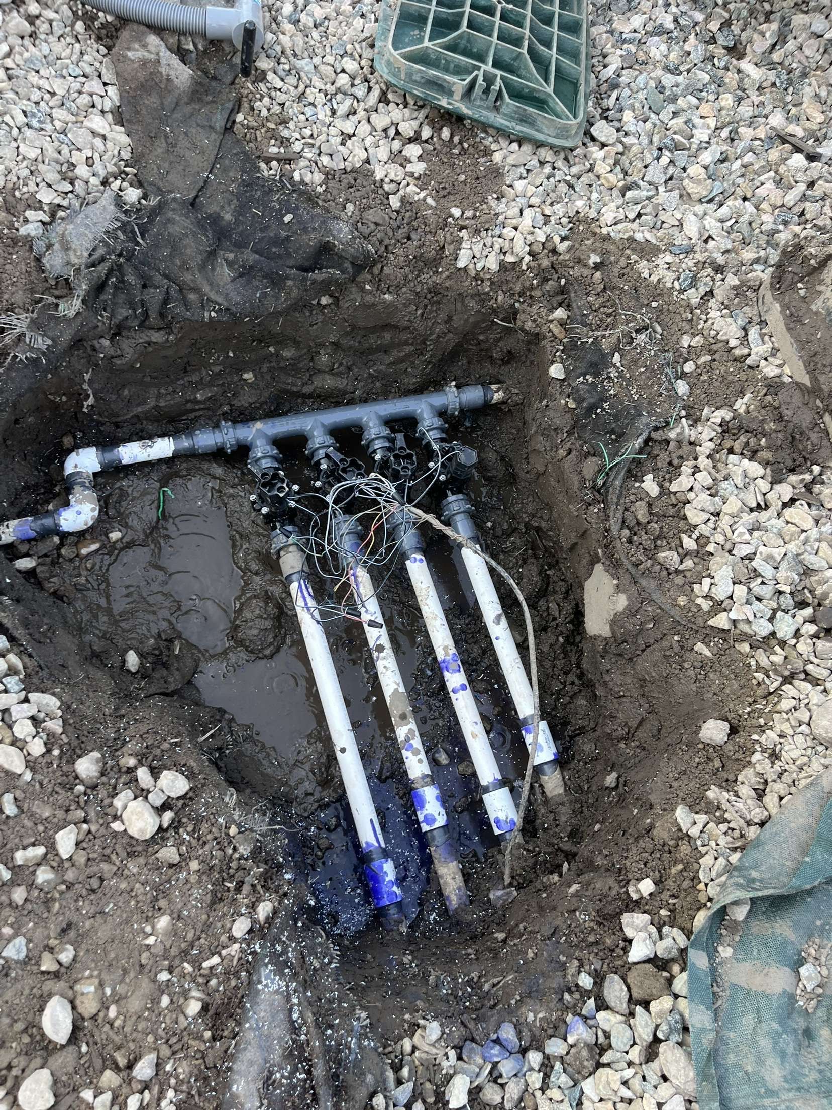 Trench showing exposed black and white utility cables in muddy soil near green plastic crates.