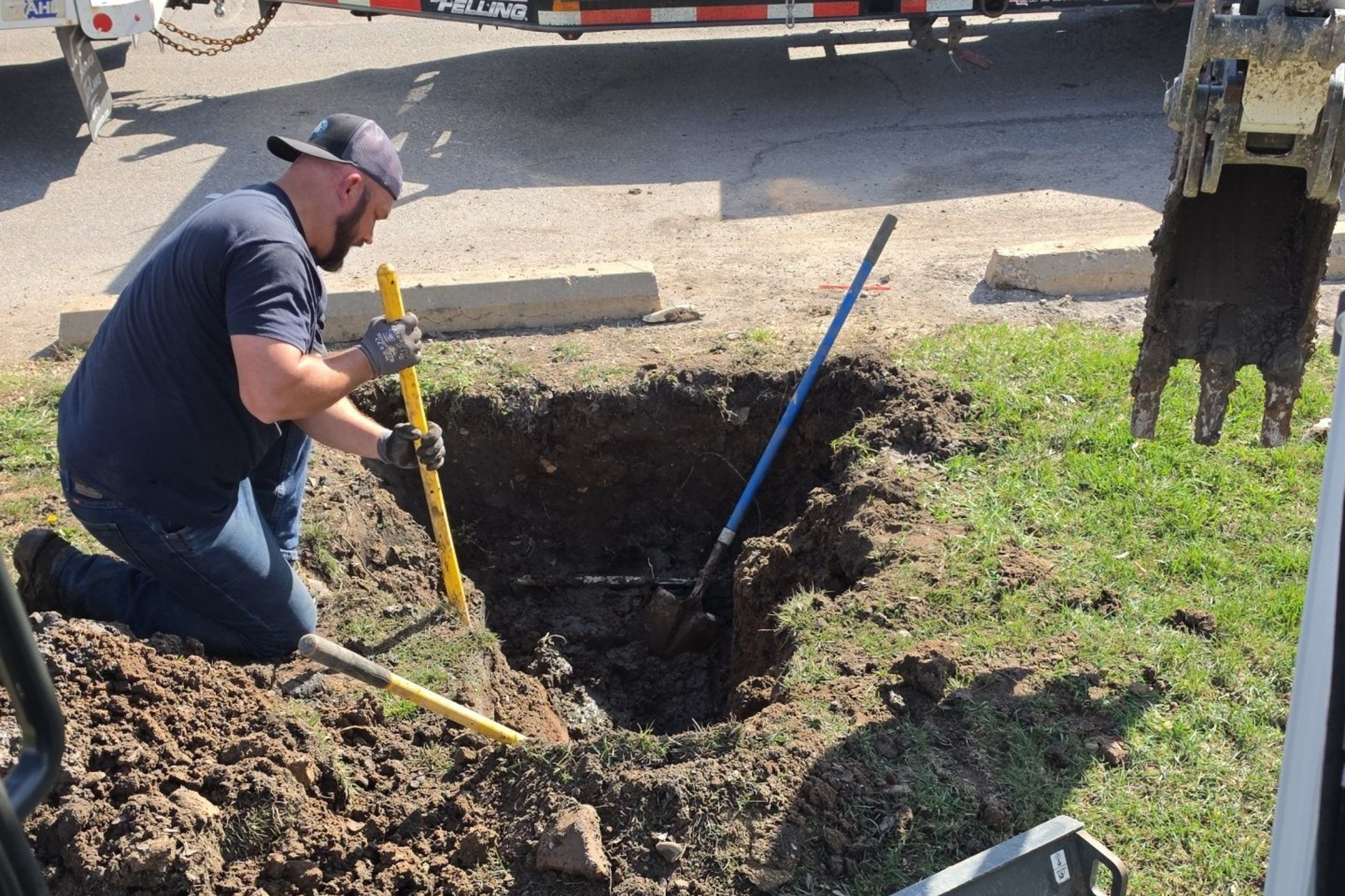 Worker digging a trench beside a yellow backhoe in a dirt yard